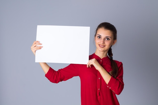 Jovem mulher agradável na blusa vermelha segurando uma folha de papel branca sobre fundo cinza no estúdio