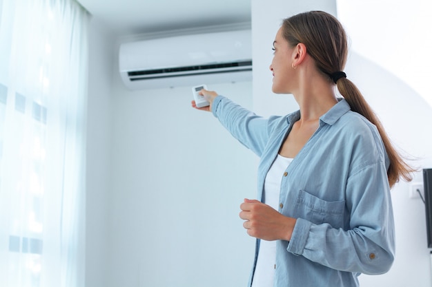 Jovem mulher ajusta a temperatura do ar condicionado usando o controle remoto no quarto em casa