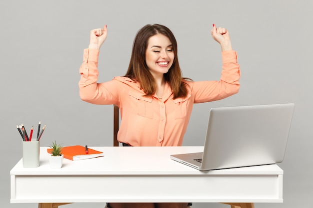 Jovem mulher alegre em roupas casuais pastel fazendo gesto de vencedor, sente-se trabalhar na mesa branca com laptop pc isolado em fundo cinza. Conceito de estilo de vida de carreira de negócios de realização. Mock up espaço de cópia.