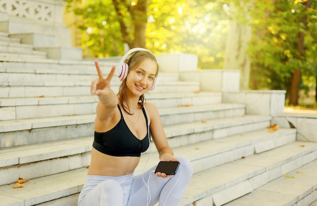 Jovem mulher alegre em roupas esportivas ouve música em fones de ouvido e mostra um gesto V enquanto está sentado na escada em um dia ensolarado.