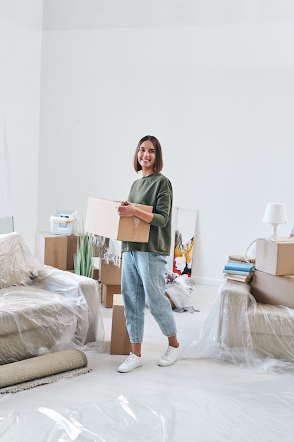 Jovem mulher alegre em trajes casuais carregando caixa com coisas enquanto se move pela sala de estar de um apartamento ou casa nova