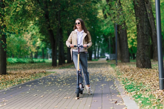 Jovem mulher andando de scooter elétrica em um parque de outono. Transporte verde, problemas de congestionamento.