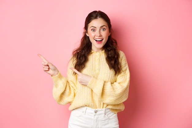 Jovem mulher animada em roupa casual, mostrando o banner, apontando os dedos para o lado esquerdo do espaço da cópia, em pé contra a parede rosa.