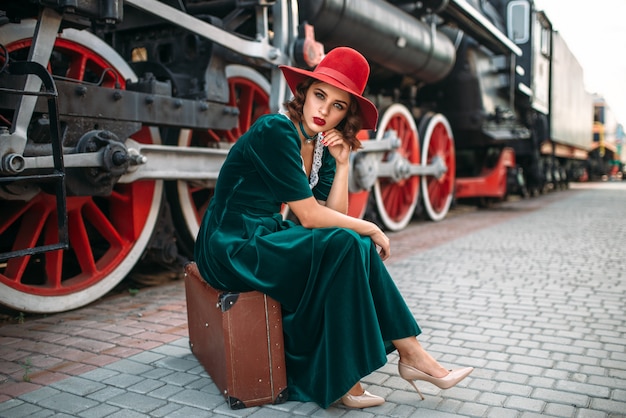 Jovem mulher antiquada sentada na mala contra o trem a vapor vintage, closeup de rodas vermelhas. Locomotiva velha. Motor ferroviário, viagem de trem
