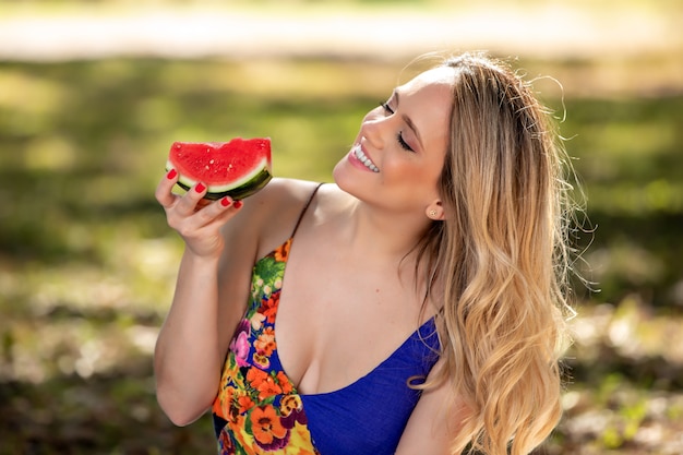 Jovem mulher ao ar livre sob as árvores em um parque, feliz e sorridente, fazendo pic nic e degustação de melancia.