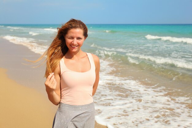 Jovem mulher apertando um olho, enquanto o sol forte brilha sobre ela na praia, mar ao fundo.