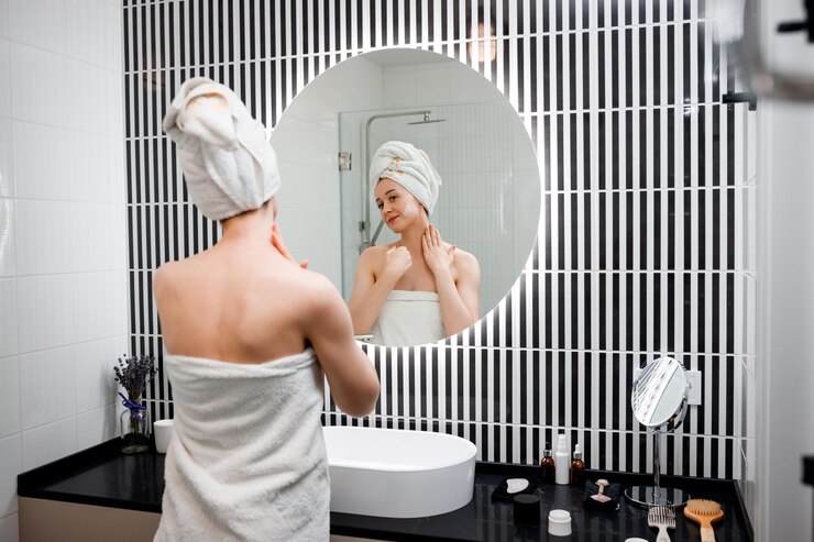 Foto jovem mulher aplicando creme antirrugas em pé atrás do espelho no banheiro doméstico cosmetologia e procedimento de beleza cuidados com a pele após a limpeza