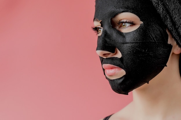 Jovem mulher aplicar máscara facial de tecido cosmético preto sobre fundo rosa. Máscara de peeling facial com carvão, tratamento de beleza spa, cuidados com a pele, cosmetologia. Fechar-se