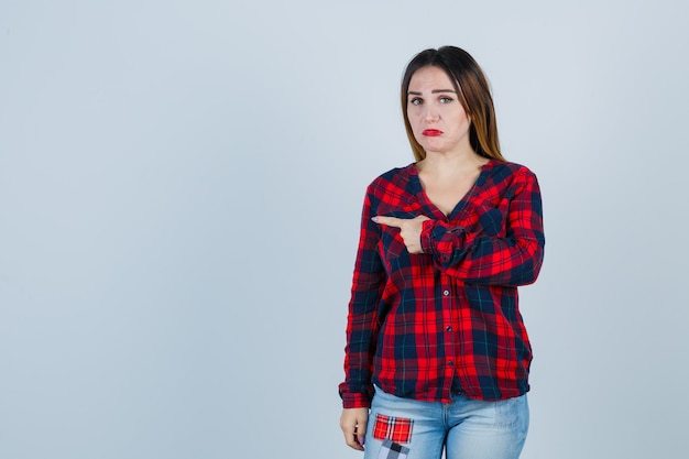 Jovem mulher apontando para a esquerda com o dedo indicador em camisa xadrez, jeans e parecendo chateada. vista frontal.