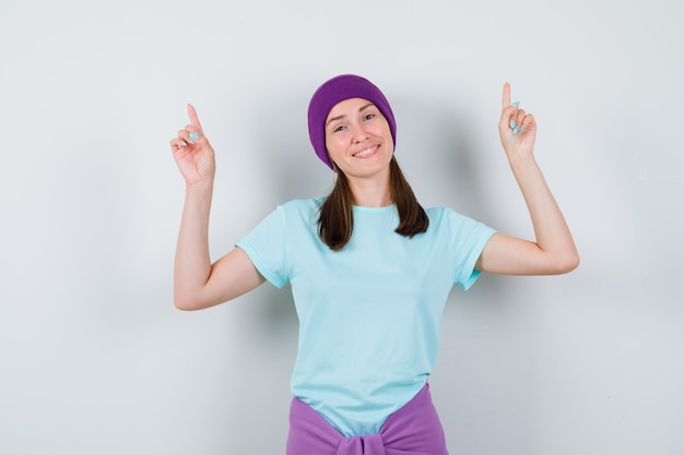 Jovem mulher apontando para cima com os dedos indicadores em t-shirt azul, gorro roxo e olhando alegre. vista frontal.