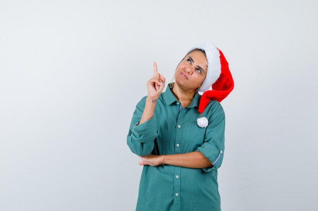 Jovem mulher apontando para cima na camisa, chapéu de Papai Noel e olhando confiante, vista frontal.