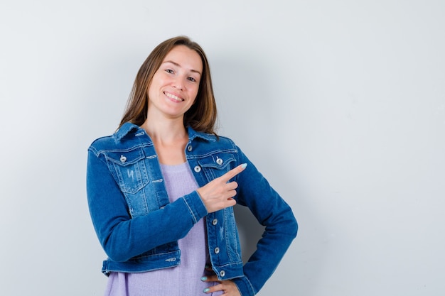 Jovem mulher apontando para o canto superior direito de camiseta, jaqueta e parecendo alegre. vista frontal.