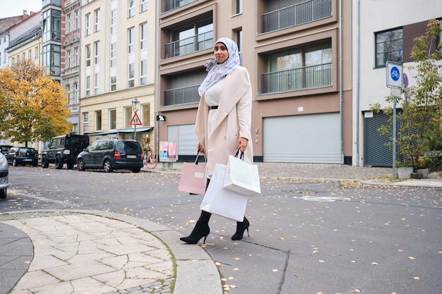 Jovem mulher árabe atraente em hijab andando pela aconchegante rua da cidade com sacolas de compras