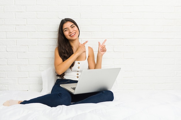 Jovem mulher árabe que trabalha com seu laptop na cama apontando com o dedo indicador para um espaço de cópia, expressando emoção e desejo.