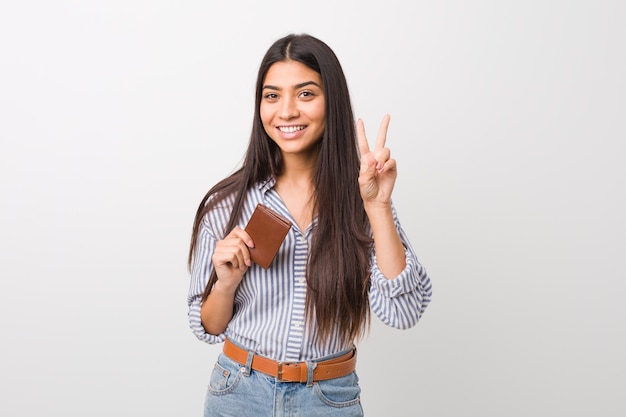 Jovem mulher árabe segurando uma carteira mostrando sinal de vitória e sorrindo amplamente.