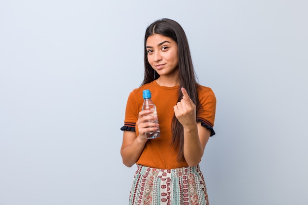 Jovem mulher árabe segurando uma garrafa de água, apontando com o dedo para você, como se estivesse convidando