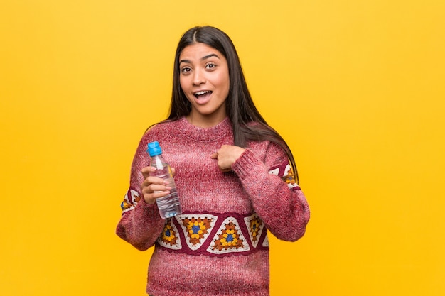 Jovem mulher árabe segurando uma garrafa de água surpreendeu apontando para si mesmo, sorrindo amplamente.