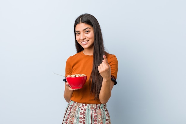 Jovem mulher árabe segurando uma tigela de cereais, apontando com o dedo para você, como se estivesse convidando para se aproximar.