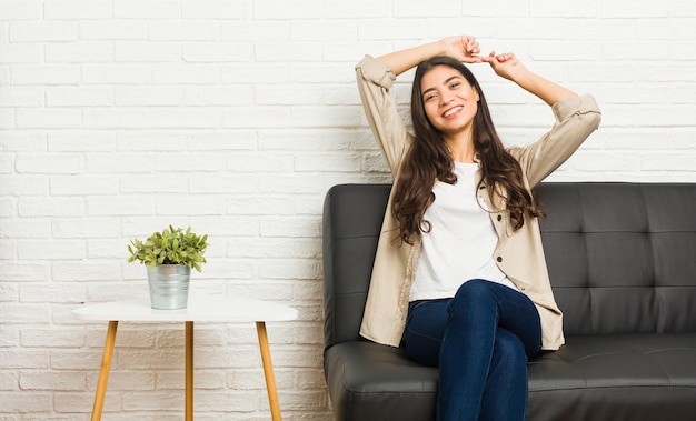 Jovem mulher árabe sentada no sofá, esticando os braços, posição relaxada.