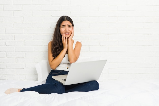 Jovem mulher árabe trabalhando com seu laptop na cama choramingando e chorando desconsolado.