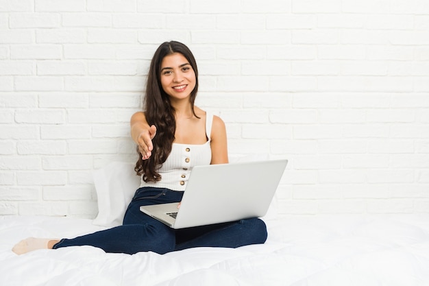 Jovem mulher árabe trabalhando com seu laptop na cama, esticando a mão para a câmera em um gesto de saudação.