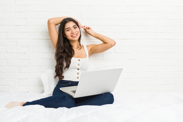 Jovem mulher árabe trabalhando com seu laptop na cama esticando os braços, posição relaxada.