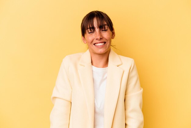 Jovem mulher argentina isolada em fundo amarelo ri e fecha os olhos, sente-se relaxada e feliz.