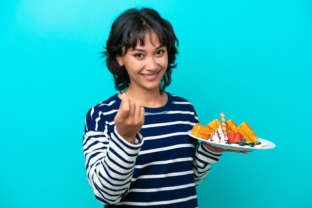 Foto jovem mulher argentina segurando waffles isolada em fundo azul fazendo gesto de dinheiro