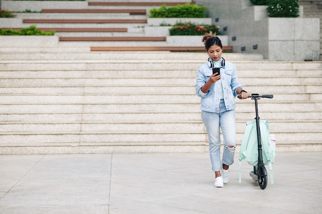 Jovem mulher asiática ao lado de sua scooter elétrica com uma mochila e lendo mensagens em seu telefone