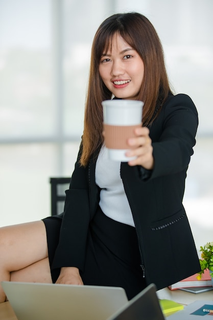Jovem mulher asiática atraente em um terno preto, segurando uma xícara de café no escritório moderno com fundo borrado do windows. Conceito de estilo de vida de escritório moderno.