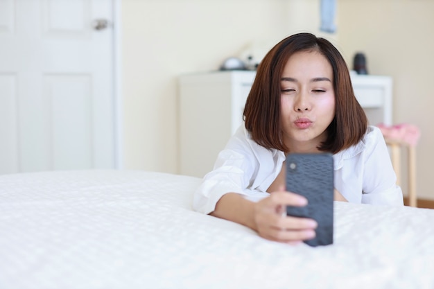 Jovem mulher asiática atraente vestindo uma camisa branca usando uma chamada VDO do celular e mandando um beijo para alguém enquanto está sentado no quarto