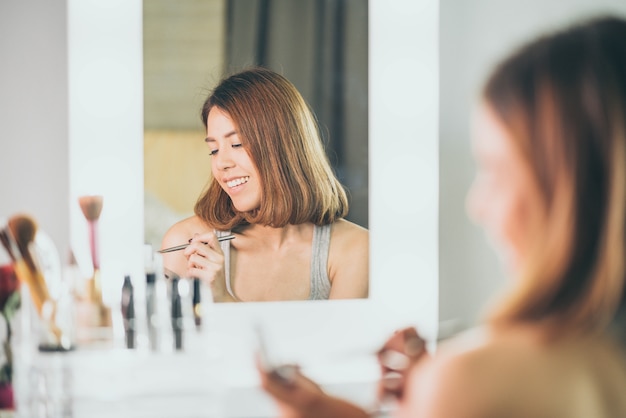 Jovem mulher asiática bonita fazendo maquiagem perto de espelho