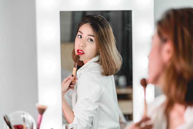 Jovem mulher asiática bonita fazendo maquiagem perto de espelho