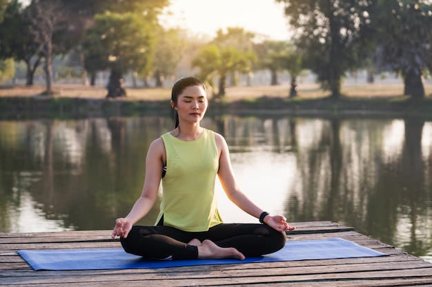 Jovem mulher asiática bonita praticando ioga e meditando na pose de lótus ao ar livre ao lado do lago pela manhã para relaxamento e paz de espírito. conceito de harmonia e meditação. estilo de vida saudável