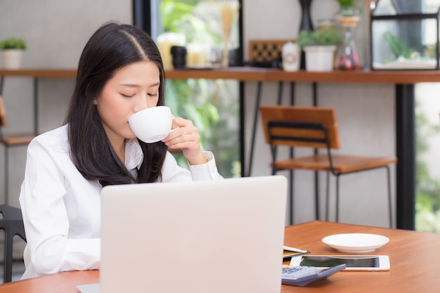 Jovem mulher asiática bonita que trabalha em linha no portátil e bebe o café.