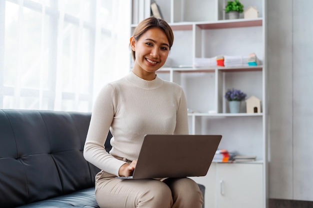 Jovem mulher asiática bonita usando laptop ao sentar no sofá na sala de estar