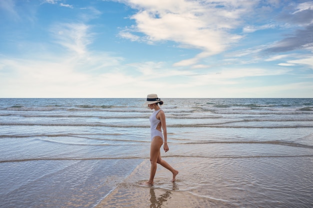 Jovem mulher asiática com chapéu em trajes de banho, caminhando relaxando na praia com o céu azul no mar tropical. conceito de verão e férias