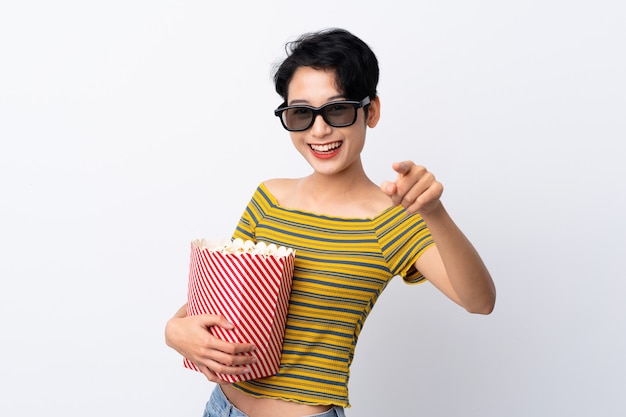 Jovem mulher asiática com óculos 3d e segurando um grande balde de pipocas enquanto aponta a frente