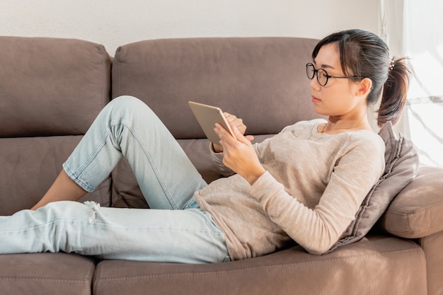 Jovem mulher asiática com óculos usando um tablet no sofá.