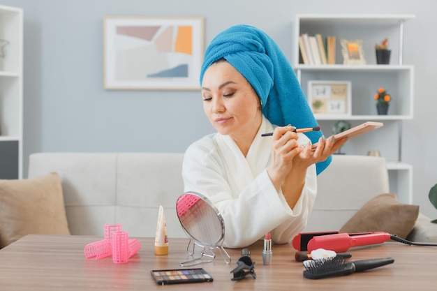 Jovem mulher asiática com toalha na cabeça sentada na penteadeira em casa interior aplicando sombras para os olhos segurando paleta fazendo beleza de rotina de maquiagem matinal e conceito de cosméticos faciais