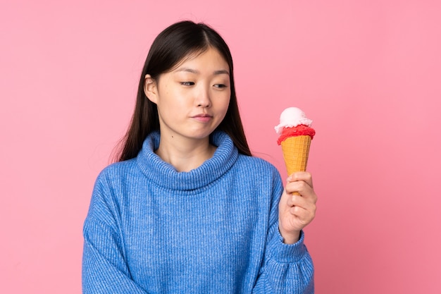 Jovem mulher asiática com um sorvete de corneta isolado na parede rosa com expressão triste