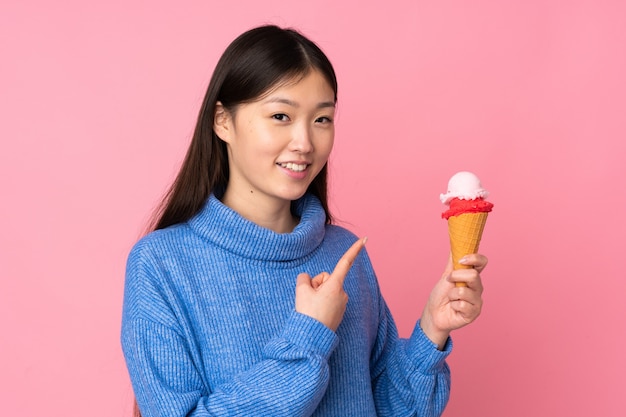 Jovem mulher asiática com um sorvete de corneta isolado no espaço rosa e apontando-o