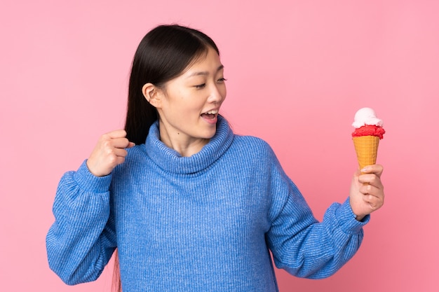 Jovem mulher asiática com um sorvete de corneta na parede rosa comemorando uma vitória