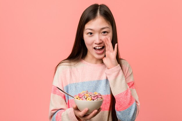 Jovem mulher asiática com uma tigela de cereal gritando animado para a frente.