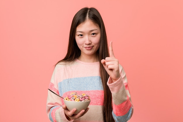 Jovem mulher asiática com uma tigela de cereal, mostrando o número um com o dedo.