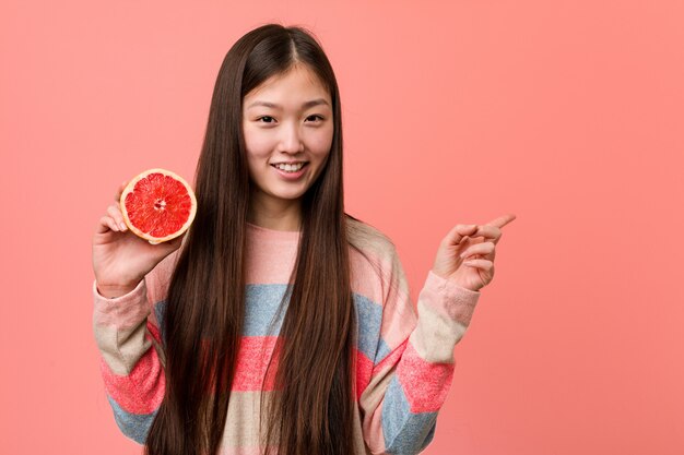 Jovem mulher asiática com uma toranja sorrindo alegremente apontando com o dedo indicador fora.