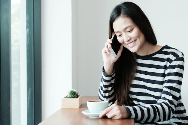 Jovem, mulher asiática, conversando com telefone inteligente, sentado ao lado da janela, café, fundo