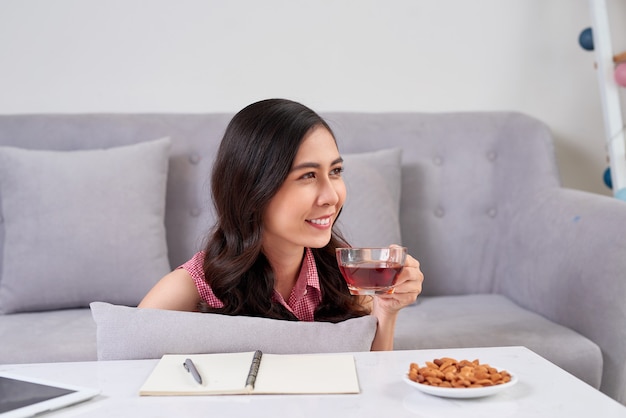 Jovem mulher asiática, desfrutando de uma pausa para o chá e um lanche, enquanto trabalhava em casa.