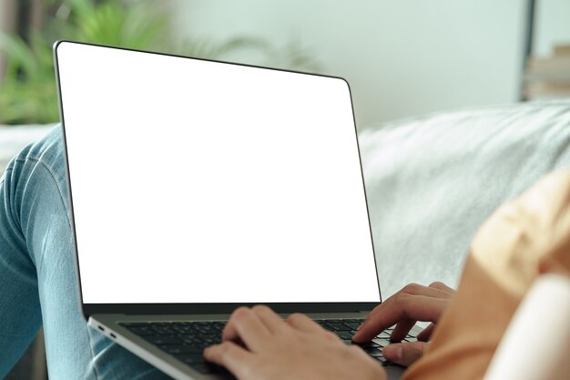 Jovem mulher asiática digitando o teclado do computador laptop com tela branca em branco no sofá. trabalhar em casa, relaxar, fazer compras online, conceito de comunicação e tecnologia
