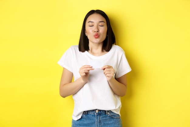 Jovem mulher asiática em camiseta branca casual mostrando cartão de crédito de plástico e lábios franzidos bobo